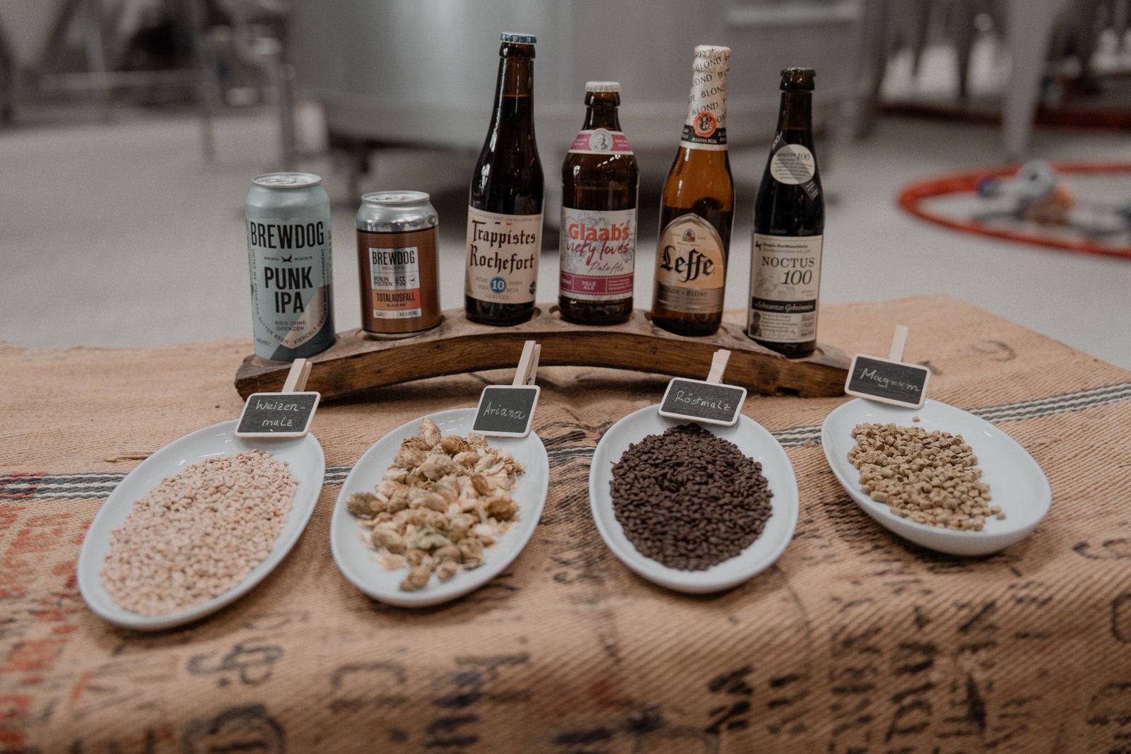 Bierrohstoffe Malz und Hopfen in Schalen neben verschiedenen Bierflaschen